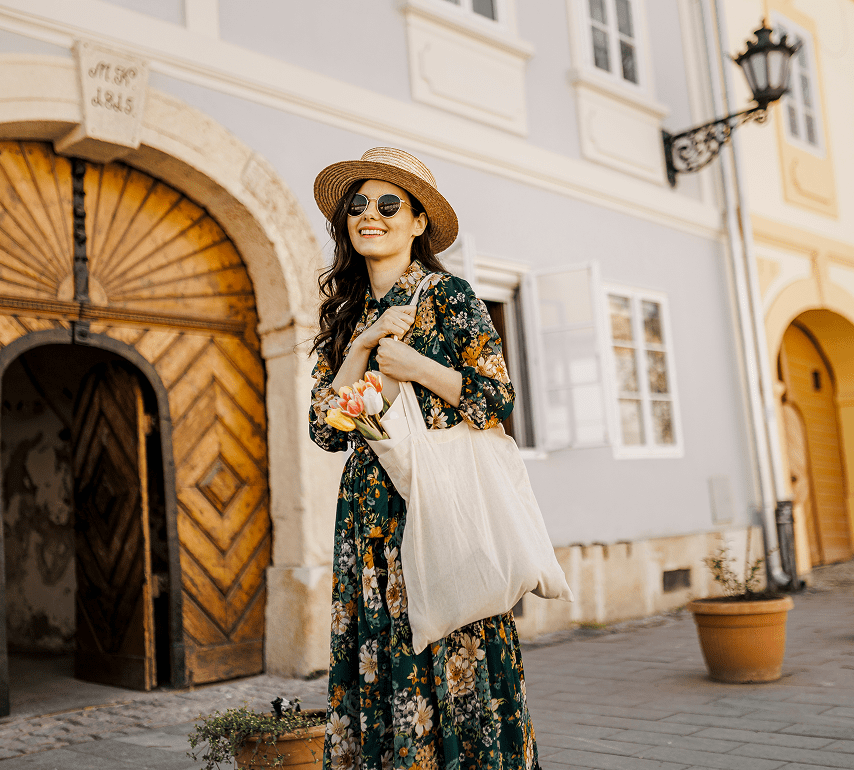 A woman in a floral dress and straw hat stands on a cobblestone street, holding a tote bag with flowers, in front of a historic building with arched wooden doors.