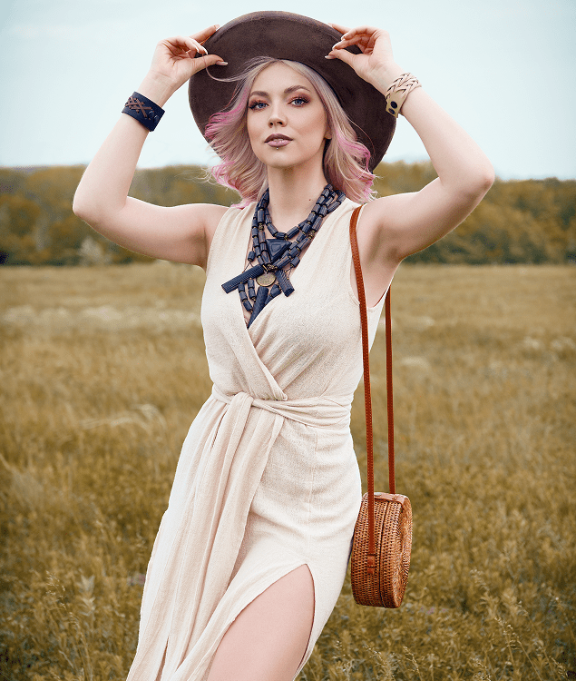 Woman in a light beige dress and wide-brim hat stands in a grassy field, holding her hat with both hands and wearing layered necklaces and a crossbody woven bag.