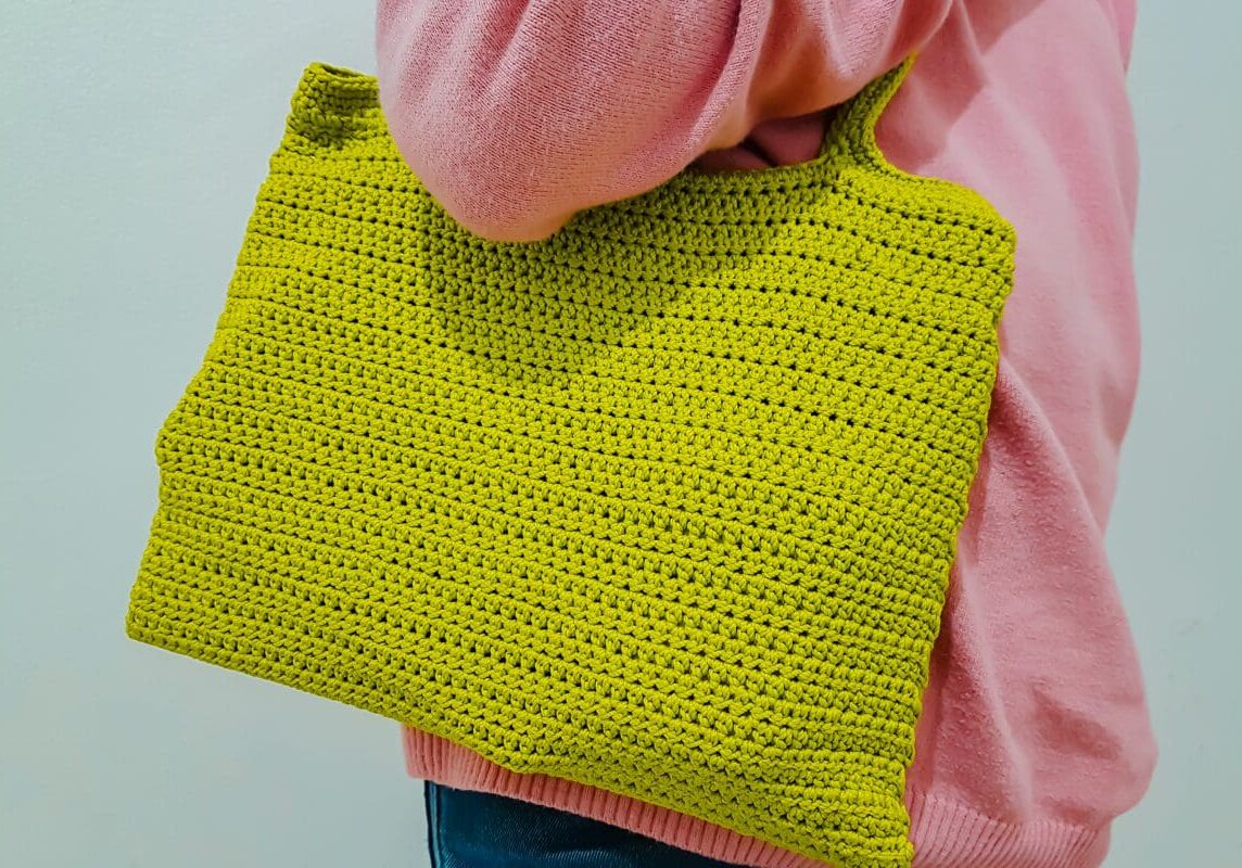 Person wearing a pink sweater holding a yellow crocheted tote bag under their arm against a plain background.
